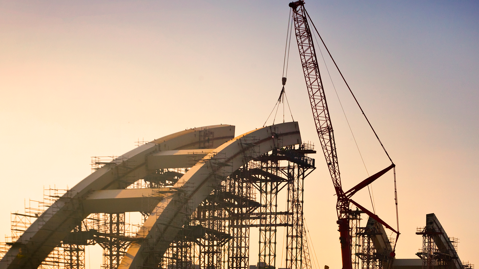 A crane lifts a steel beam onto an arched bridge as project managers oversee progress with construction IoT at sunset.