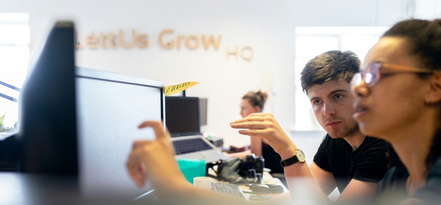 Two people discuss something on a computer in a modern office labeled Let Us Grow HQ, highlighting a connected jobsite.