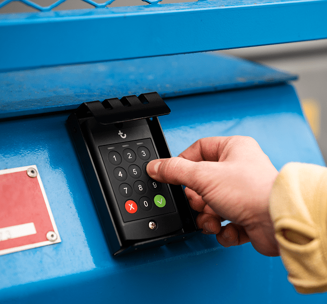 A person’s hand presses a button on a black keypad attached to blue metal, likely entering a code for connected jobsite access.