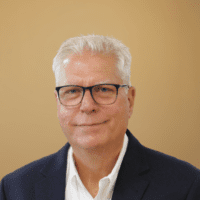 A man with short white hair and glasses in a dark blazer smiles in front of a beige background, suggesting construction IoT expertise.