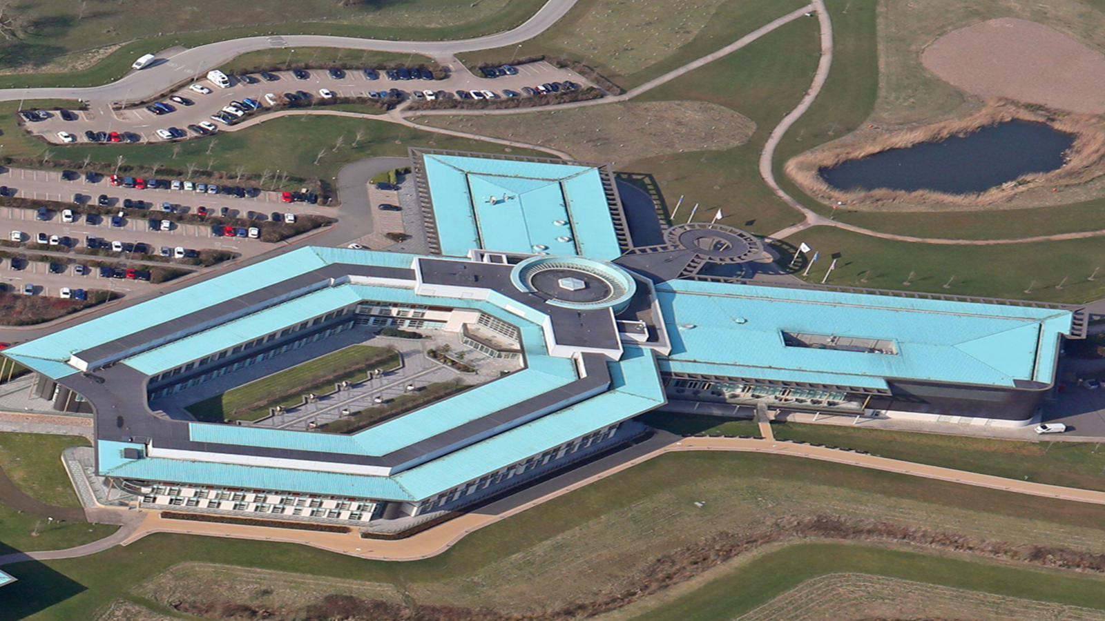 Aerial view of a modern building with a blue roof and geometric design, surrounded by green lawns, parking lots, and a small pond; ideal for connected jobsite.