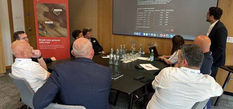 Eight people at a conference table watch a screen presentation on construction equipment tracking; one stands and gestures to the screen.