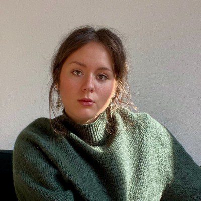 A woman with long brown hair sits indoors in a green knit sweater, looking calmly at the camera, sunlight casting soft shadows on a plain white wall behind her. She appears to be reflecting on her experience at the Downtime Summit NYC focused on construction equipment tracking.