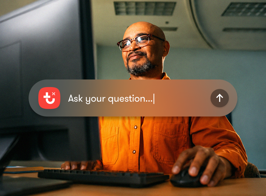 A man in glasses and an orange shirt sits at a computer with a digital search bar, suggesting a connected jobsite setting.