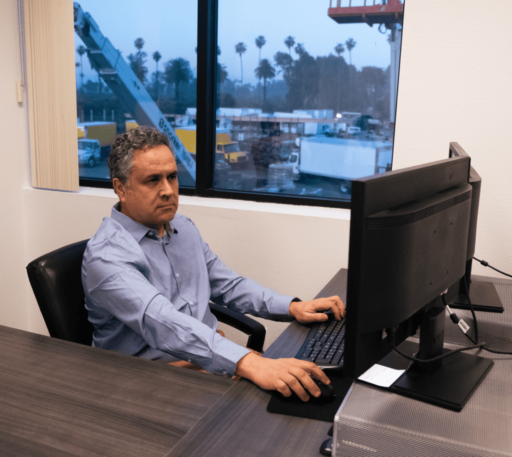 A man in a blue shirt works at a desk with dual monitors in an office overlooking a construction site, showing connected jobsite.