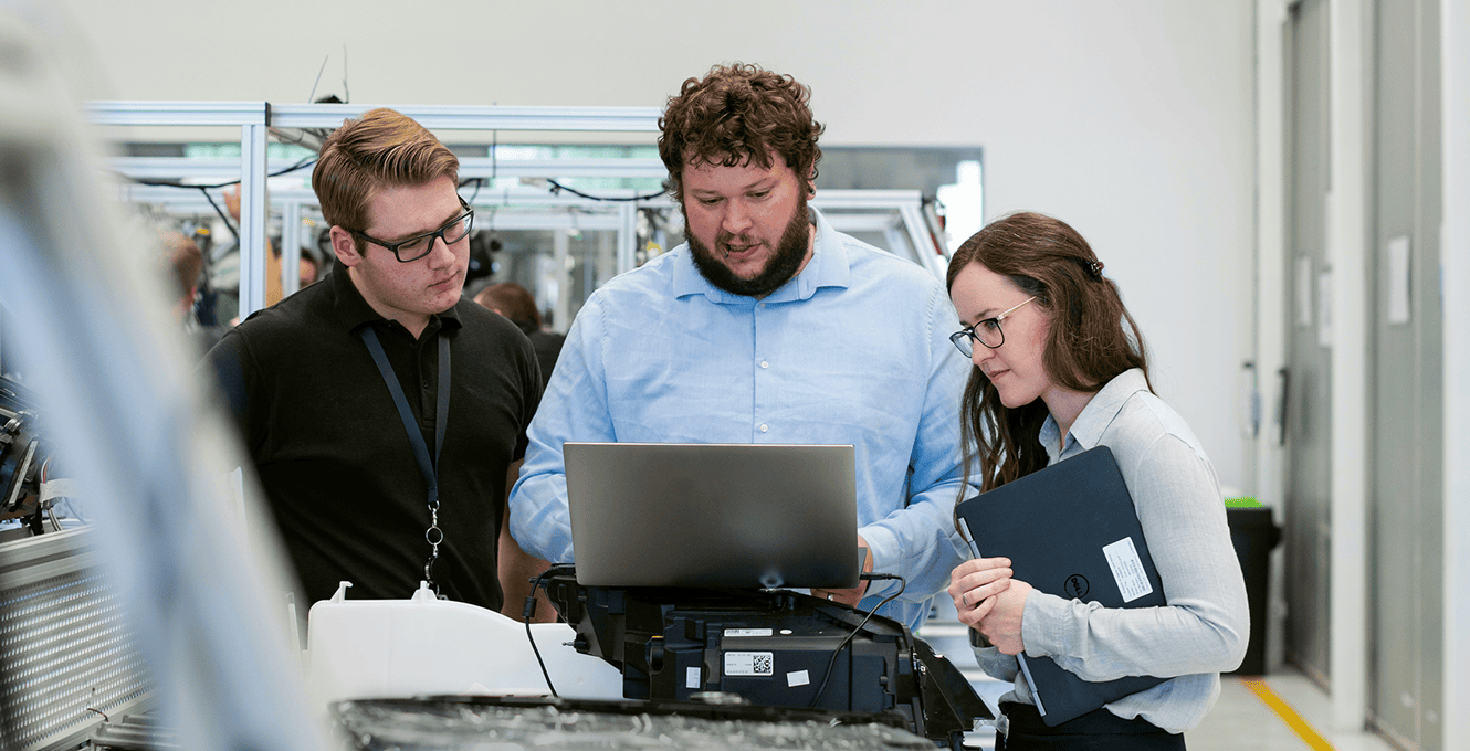 Three people stand together in a modern lab, closely examining a laptop while surrounded by equipment, suggesting a collaborative discussion about fleet management or telematics data.