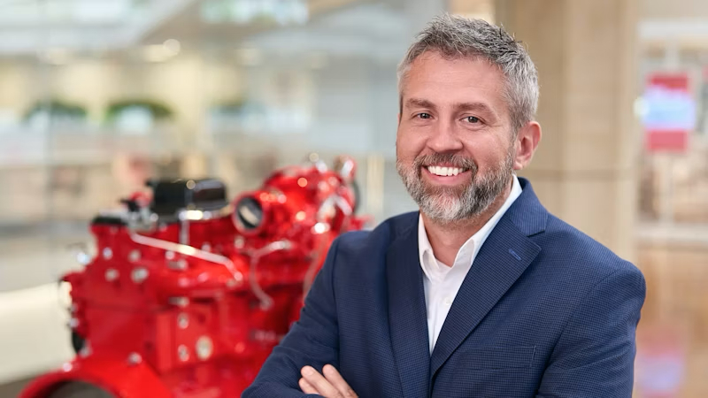 A smiling man with gray hair and beard in a blue suit stands in front of a red industrial machine with the latest IrisX technology, highlighting construction equipment tracking in a modern indoor setting.