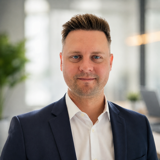 A man in a navy suit and white shirt stands in a modern office with blurred greenery and office furniture in the background, possibly considering construction equipment tracking after the Downtime Summit UK.