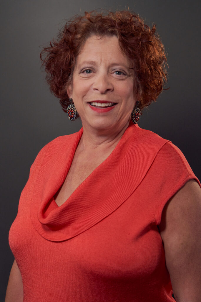 Smiling woman with curly, reddish-brown hair in a sleeveless coral top and colorful earrings stands against a dark background at the Downtime Summit NYC, related to construction equipment tracking.