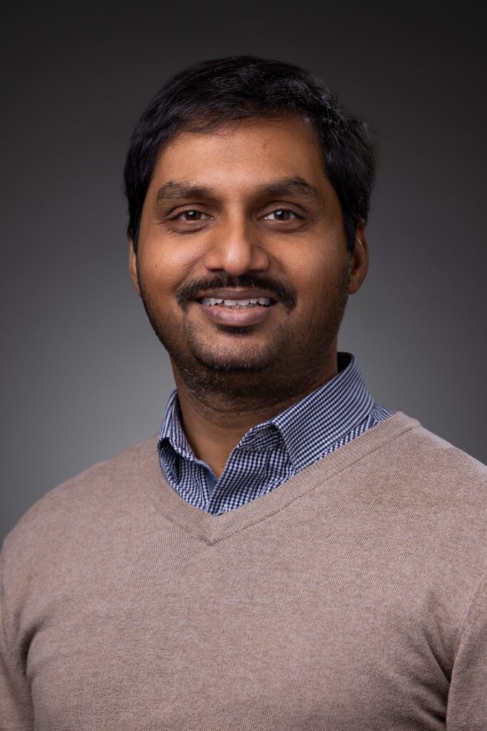 A man with short black hair and a beard smiles at the camera, wearing a light brown sweater over a blue and white checkered shirt against a plain dark background, giving the image a polished, professional feel suited for connected jobsite profiles.