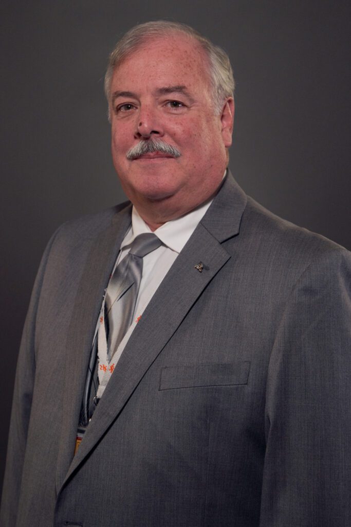 An older man with gray hair and a mustache, dressed in a light gray suit and striped tie, stands against a plain dark background, prepared for the Downtime Summit NYC focused on construction equipment tracking.