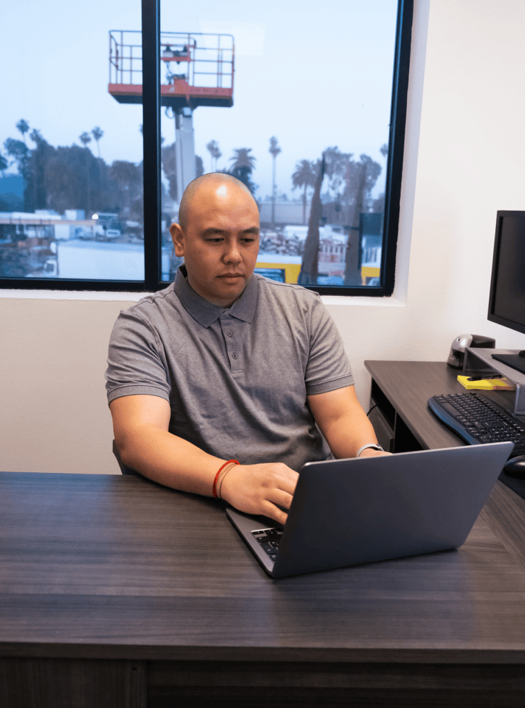 A person at an office desk works on a laptop tracking equipment emissions, with a keyboard, mouse, and monitor nearby, and construction equipment visible through a window—highlighting construction equipment tracking.