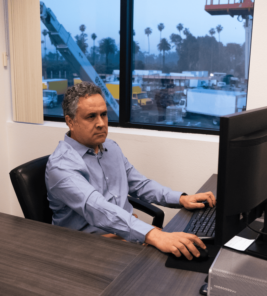 A man in a blue shirt works at a computer in an office overlooking an industrial yard with palm trees, suggesting a connected jobsite.