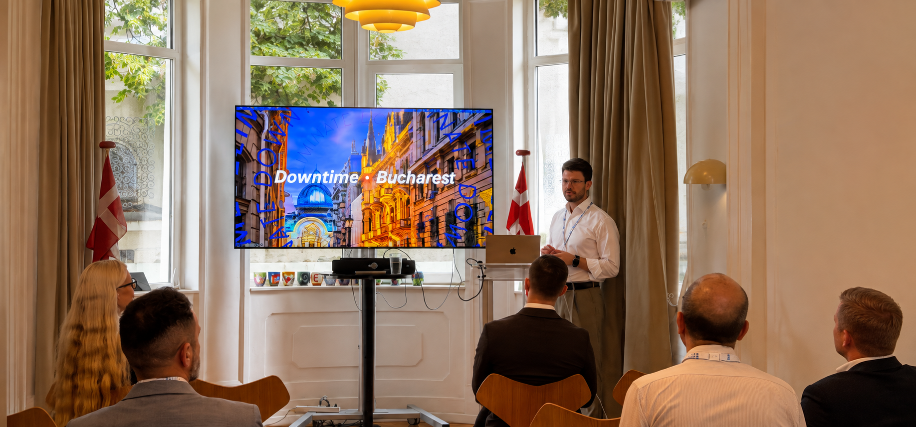 A man presents “Downtime: Bucharest” to a small audience in a bright room with large windows and Danish flags, with a screen displaying a colorful city image, relating to connected jobsite.