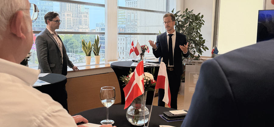 Two men in suits talk at an indoor event with Danish and Polish flags on a table, while people listen in the foreground. Large windows reveal city buildings, suggesting a connected jobsite discussion.