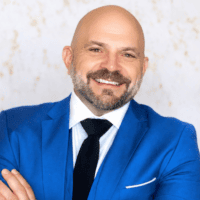 A smiling bald man with a beard, dressed in a bright blue suit, stands with arms crossed against a softly speckled background, suggesting a professional in construction equipment tracking.
