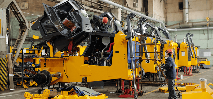 Workers in safety gear assemble and inspect large yellow industrial machinery inside a spacious factory with visible machine parts and tools, supported by an OEM data platform strategy that streamlines production and enables construction equipment tracking.