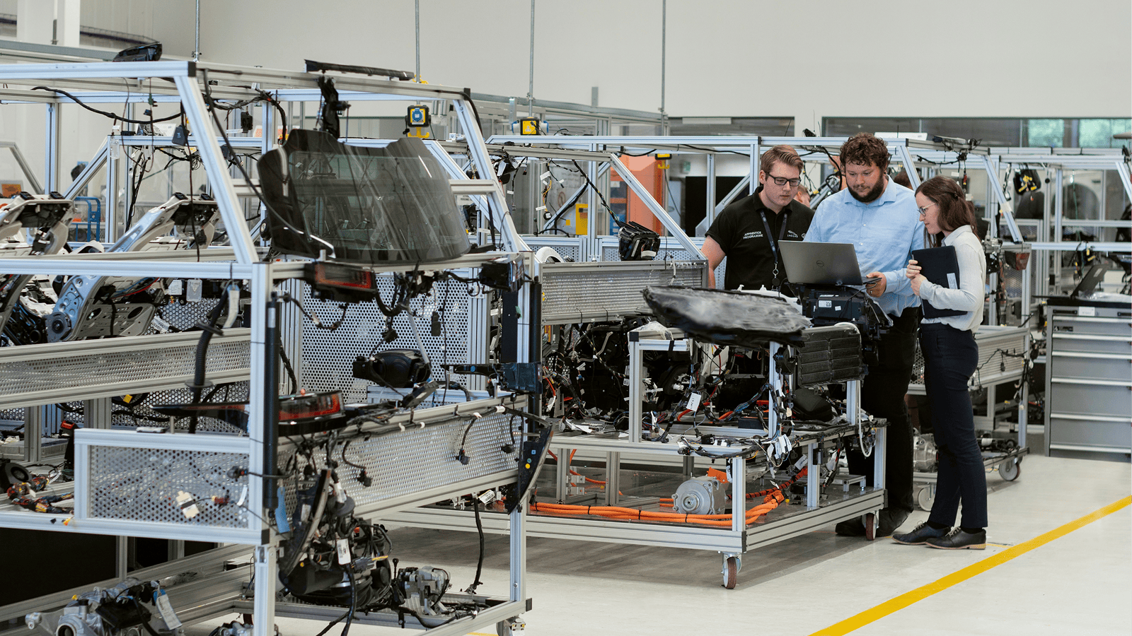 Three people review information on a laptop by a car assembly line in a modern factory, discussing their OEM data platform strategy, illustrating fleet management.