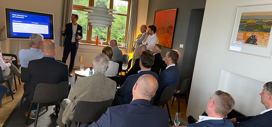 A man in a suit presents slides to an attentive group in a bright, modern room with artwork and large windows, discussing connected jobsite solutions.