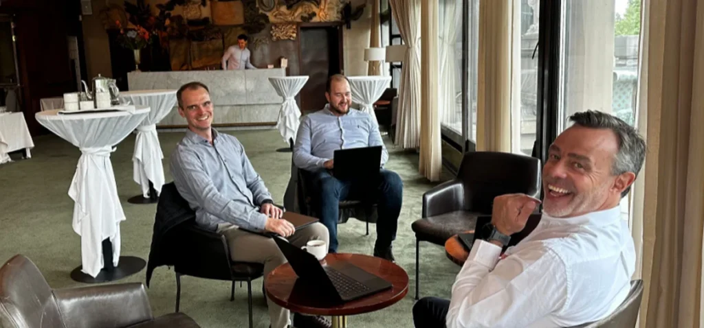 Three men in business casual attire smile while working on laptops in a hotel lounge, discussing AI on a connected jobsite in the Nordics.
