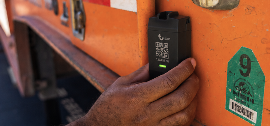 A person’s hand attaches a black asset tracking device with a QR code and green light to an orange truck or container near a green inspection sticker, illustrating construction equipment tracking.