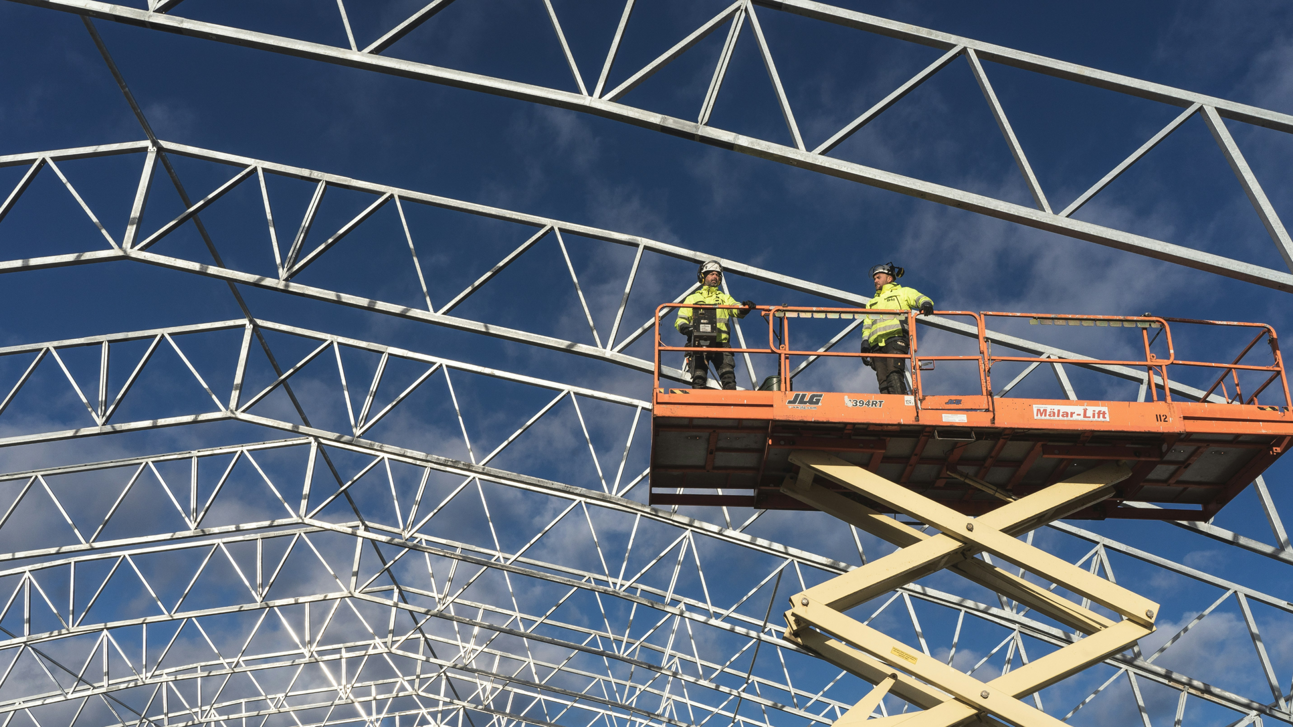 Two construction workers in safety gear stand on a scissor lift, working on the metal framework of a building under a clear blue sky, illustrating a connected jobsite.