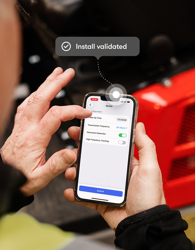 Person holding a smartphone, adjusting device settings with toggles for transmission and movement detection, as an “Install validated” message appears. Red construction equipment is visible in the background, suggesting construction IoT.