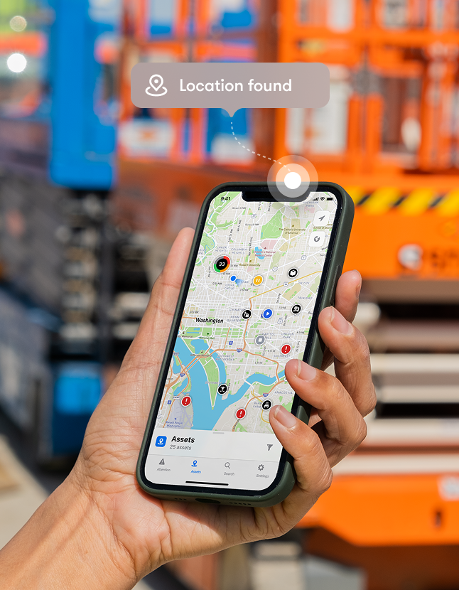 A hand holds a smartphone showing a map app with colored markers and a Location found notification, with blurred construction equipment in the background, highlighting construction equipment tracking.