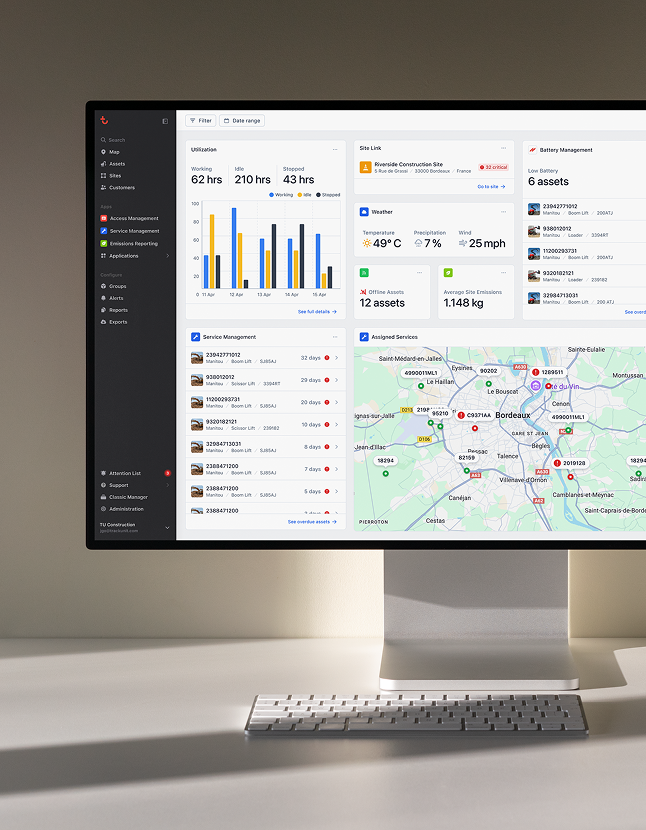 A monitor shows a fleet management dashboard with charts, stats, a device list, and a Bordeaux map displaying asset locations. A wireless keyboard rests on the white desk below.