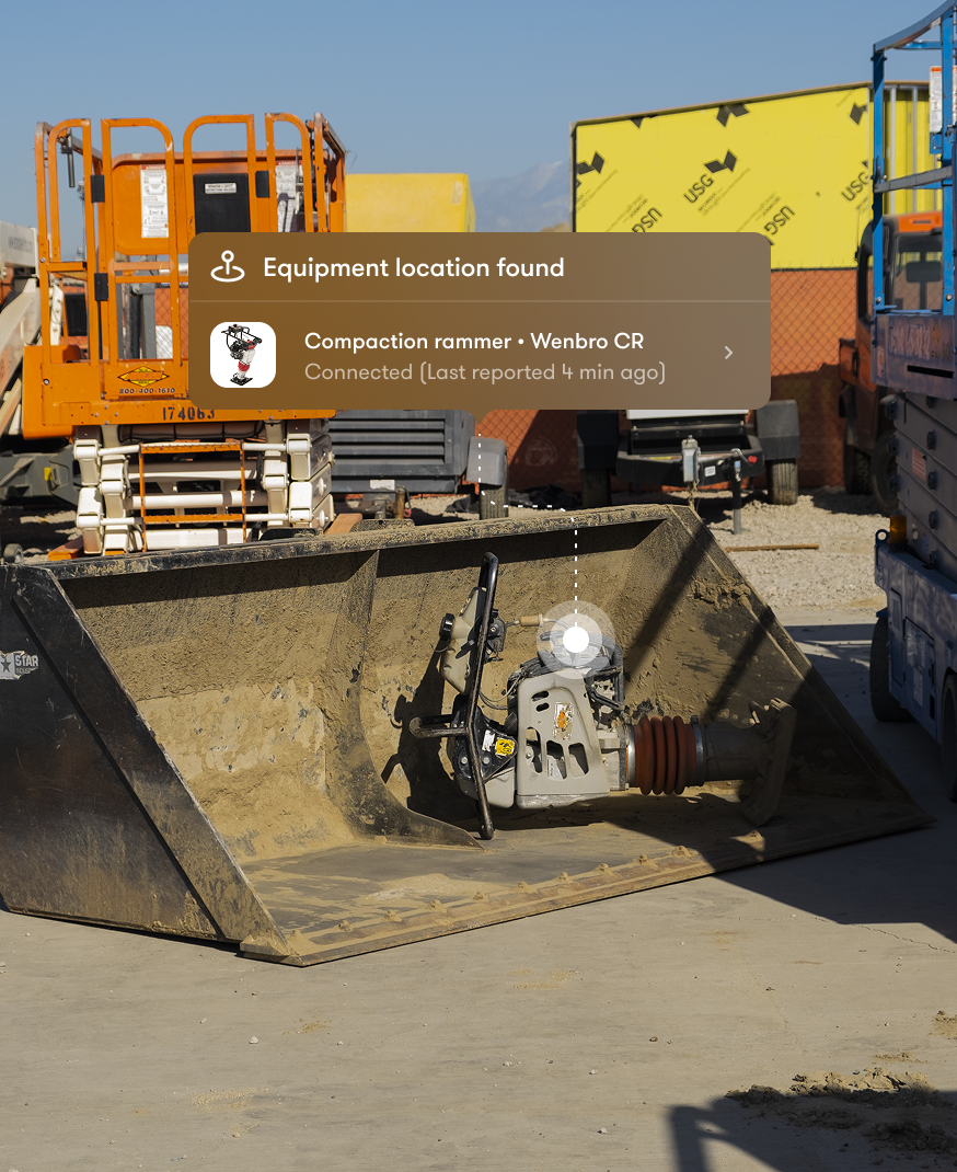A compaction rammer sits inside a large metal bucket on a construction site, with an on-screen notification showing its location and Connected status for construction equipment tracking.