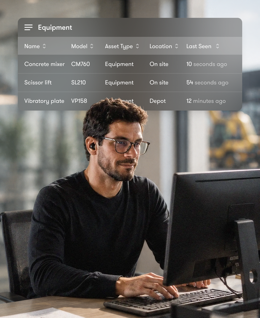 A man with glasses works at a computer while a digital overlay displays an equipment tracking table. Construction vehicles are visible through the window, highlighting construction equipment tracking.