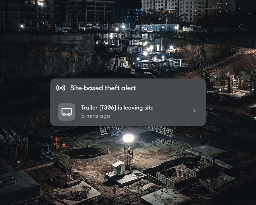 A construction site at night is lit by floodlights, with an onscreen notification about a trailer leaving the site, highlighting connected jobsite security.
