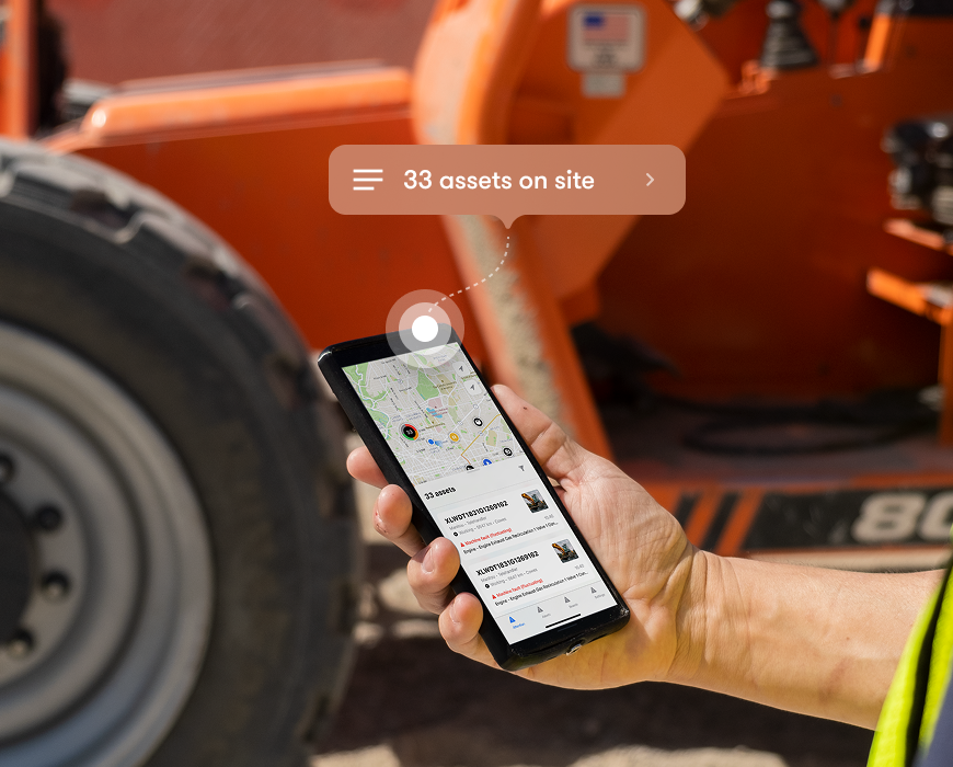 A person holds a smartphone showing a map and asset list with a graphic for 33 assets on site in front of construction equipment, illustrating construction equipment tracking.