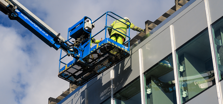 A worker in a high-visibility suit stands on a blue cherry picker, inspecting upper windows of a modern glass building under partly cloudy skies, illustrating construction equipment tracking.