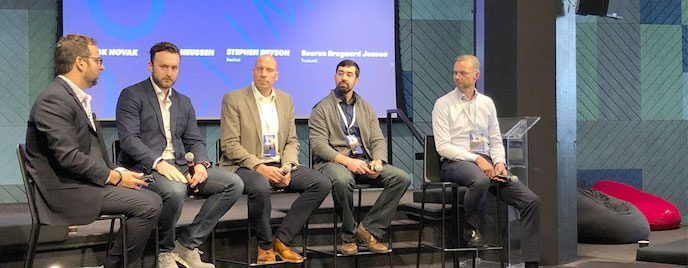 Five men sitting on high stools on a stage during a connected jobsite panel discussion, with their names displayed on the screen behind them.