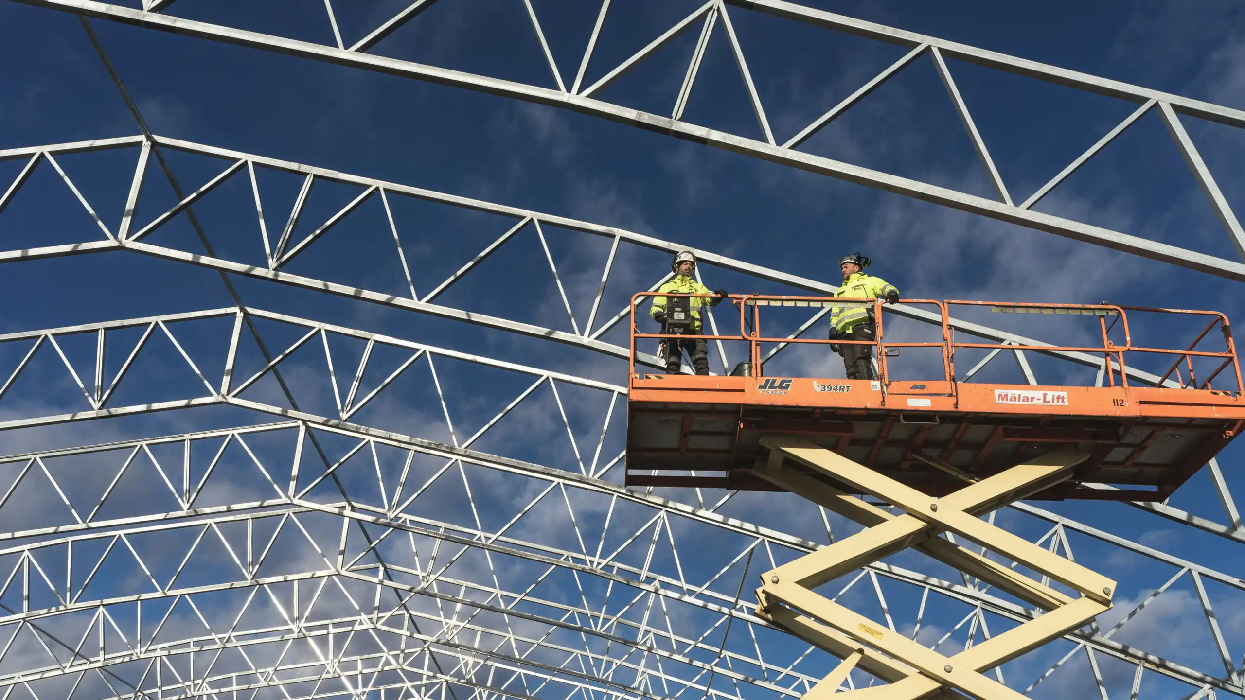 Two workers in high-visibility vests use a MEWP platform to inspect a building’s metal framework on a connected jobsite.