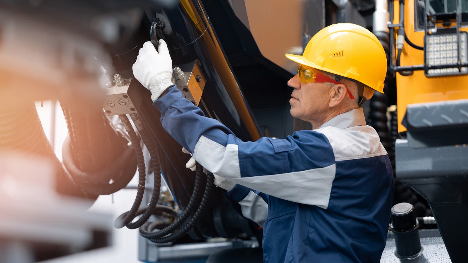A technician in a yellow hard hat and safety glasses performs maintenance on heavy construction equipment using an OEM component on a connected jobsite.