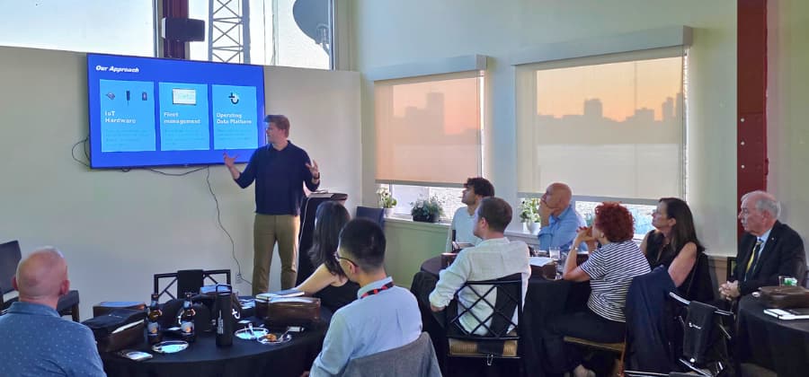 A speaker presents to a small audience in a conference room, with a screen displaying a fleet management diagram.