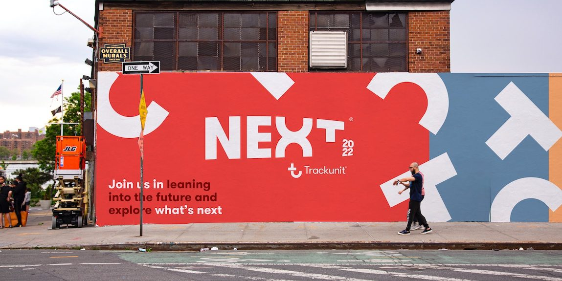 A large red mural on a construction site fence featuring NEXT 2022 and the Trackunit logo, highlighting why eliminating downtime depends on trust for a connected jobsite.