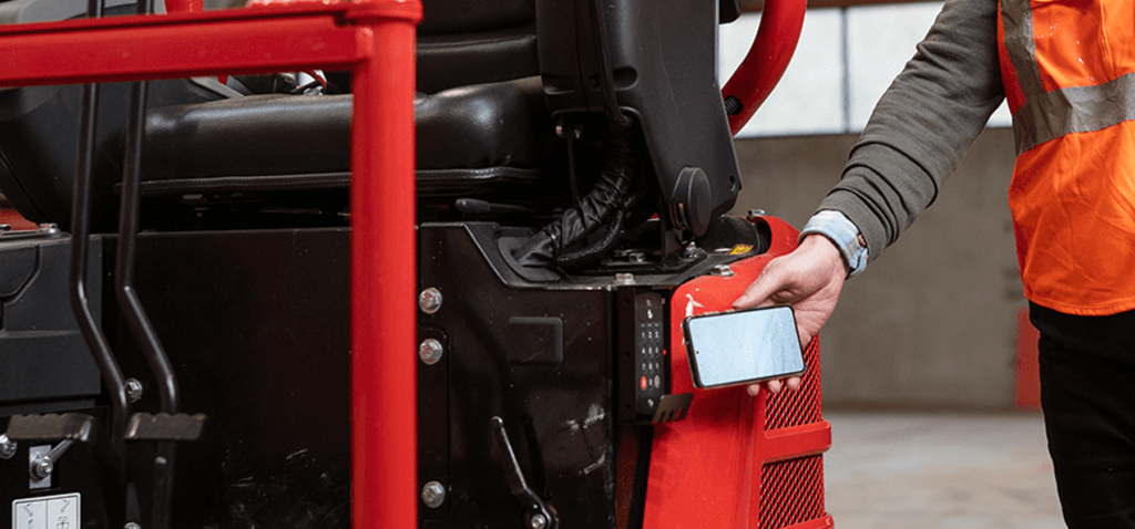 A worker in an orange high-visibility vest uses a smartphone to wirelessly access the keypad on heavy construction machinery, illustrating construction IoT.