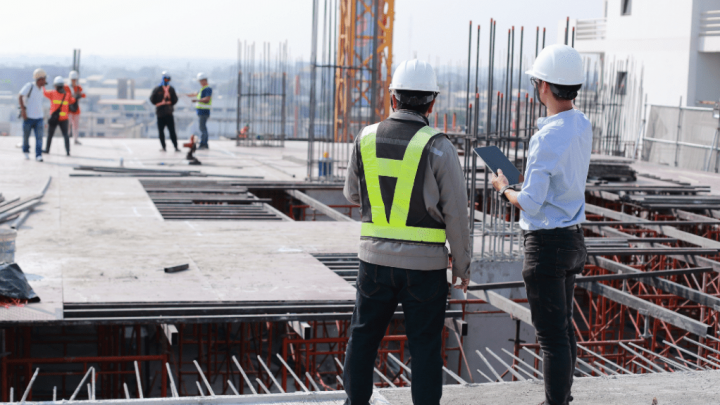 Two construction workers in safety gear discuss site plans on a building site, with others working above, showing a connected jobsite.