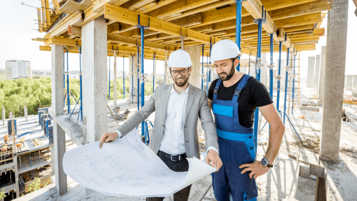 Two men in hard hats, one in a suit and one in blue overalls, review blueprints at a construction site with exposed beams—construction.