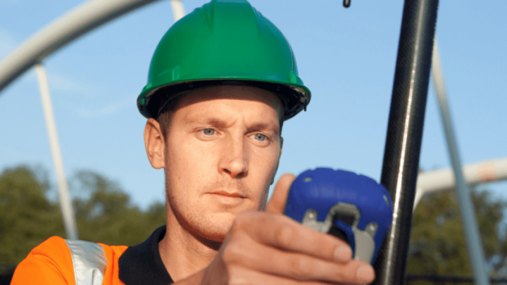 A construction worker in a green hard hat and orange vest uses a handheld device outdoors, checking for gps jammers on a connected jobsite.