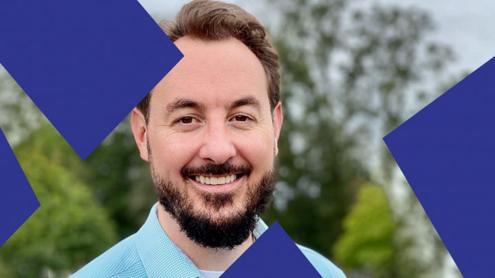 A bearded man in a light blue shirt smiles outdoors, with blurred greenery and blue shapes at an equipment rental or connected jobsite.