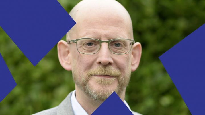 A bald man with glasses and a beard stands outdoors by greenery, with blue IoT-like shapes over the image, suggesting construction IoT.