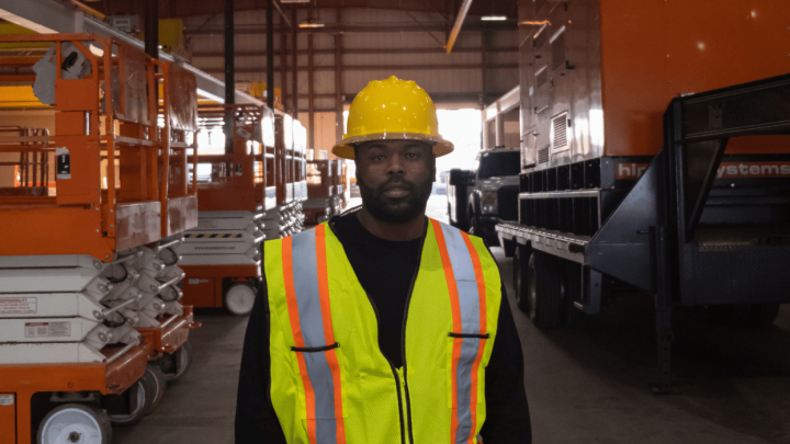 A fleet manager in a yellow hard hat and safety vest stands in a warehouse with orange construction equipment, showing fleet management.