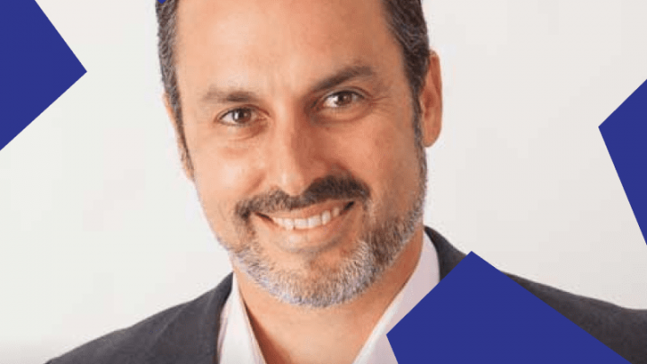 A smiling man in a white shirt and blazer stands against a light background with blue geometric shapes, suggesting construction IoT.