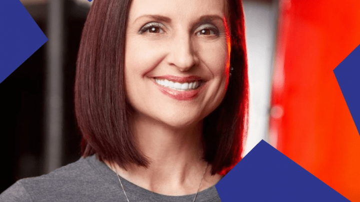 Smiling woman with shoulder-length brown hair in a gray t-shirt, standing before blue geometric shapes, representing connected jobsite.