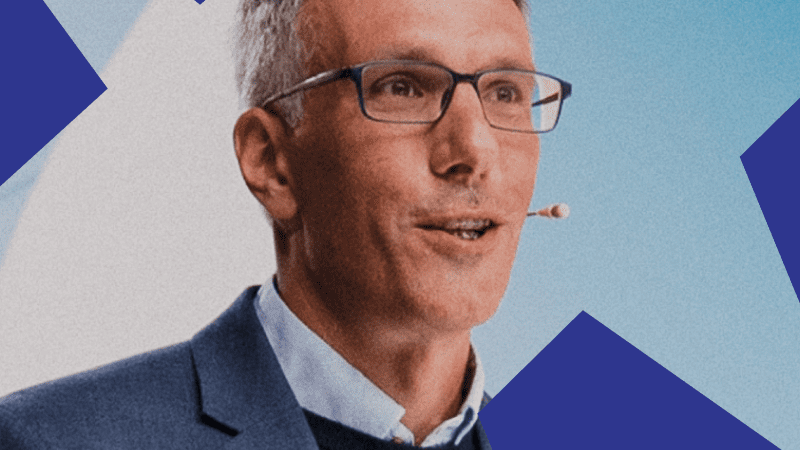 A man with short gray hair and glasses, in a suit and headset mic, speaks on stage before a blue background at a connected jobsite event.