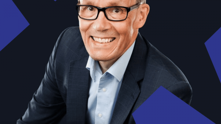 A man in a dark suit and glasses smiles at the camera; blue geometric shapes behind him suggest a connected jobsite setting.
